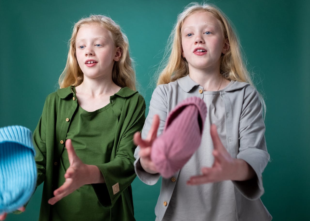 Twin girls wearing casual outfits, playfully tossing hats indoors against a green background.